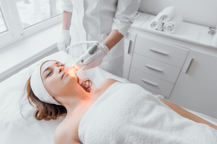 The beautician conducts light therapy of the face in a beauty clinic using an LED device. The procedure improves microcirculation and stimulates collagen production, and increases skin tone and elasticity. High quality photo
